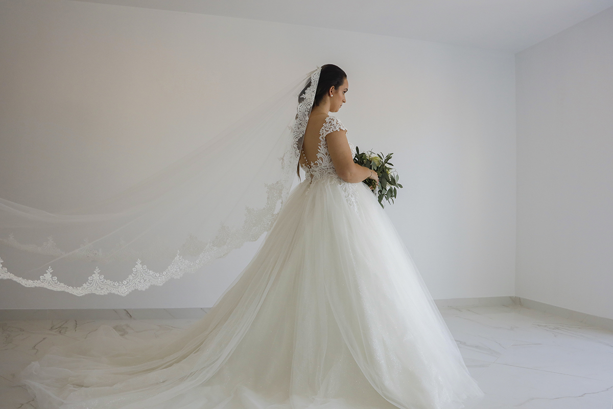 Num cenário de brancura luminosa ela surge de costas, ligeiramente de lado, segurando o bouquet à sua frente, com o véu e o vestido a arrastarem-se pelo chão numa cena de puro encanto.