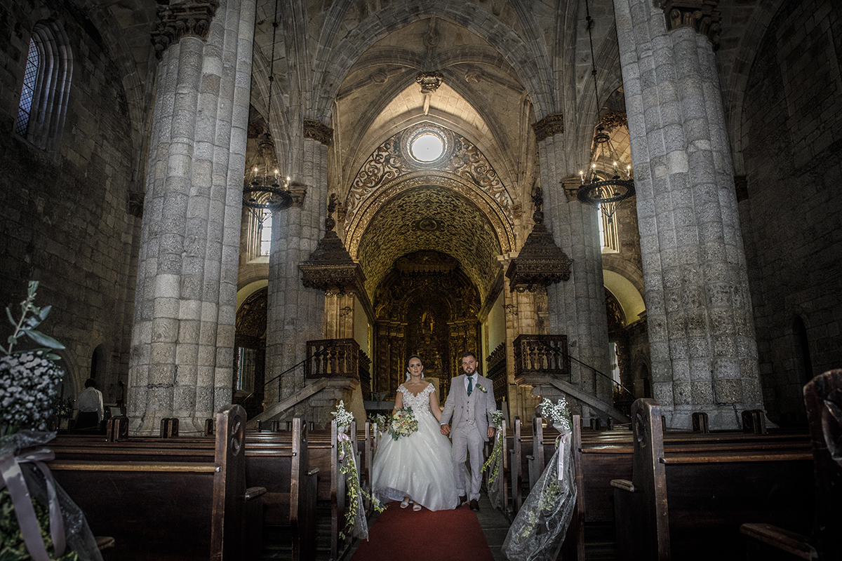 Agora virados para a porta da igreja, os noivos caminham juntos pela passadeira de mãos dadas, com os rostos iluminados pela luz da entrada e os corações a baterem em uníssono.