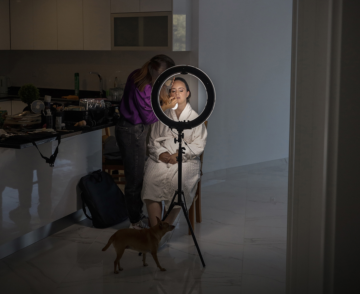 Através do círculo da ring light surge o rosto de Marta enquanto é maquilhada, um instante captado com graça e mistério onde os olhos brilham de serenidade.