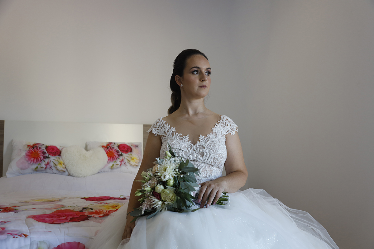 Ainda sentada, ela apoia uma mão na cama e com a outra segura o bouquet sobre as pernas enquanto olha fixamente para o lado, iluminada por uma janela que parece entender-lhe os pensamentos.