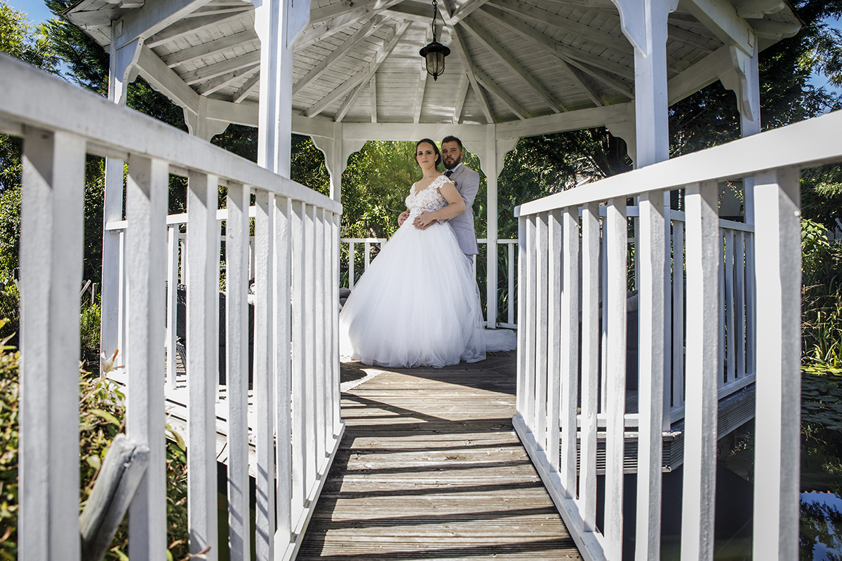 Descrição: Aqui vemos por entre um corredor de uma ponte no exterior com corrimões brancos e os dois estão ao fundo na mesma posição da foto anterior só que agora a olharem para a câmara e a sorrirem.