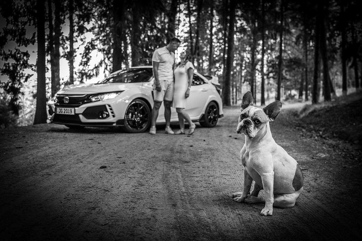 Com um toque de vinheta, esta imagem a preto e branco mostra Jorge e Sabrina no fundo, encostados ao carro, a olharem-se com amor. À frente, o cão está sentado e muito sério, a encarar diretamente a lente da câmara. Fotografia Solteiros