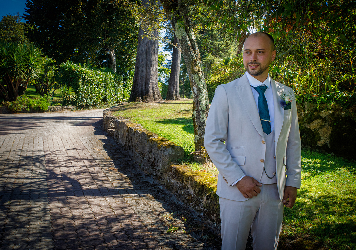 Em pé, à direita da imagem, o noivo sorri com calma. O cenário natural atrás dele, muro, erva e árvores, ecoa a simplicidade de quem encontra paz no momento presente.