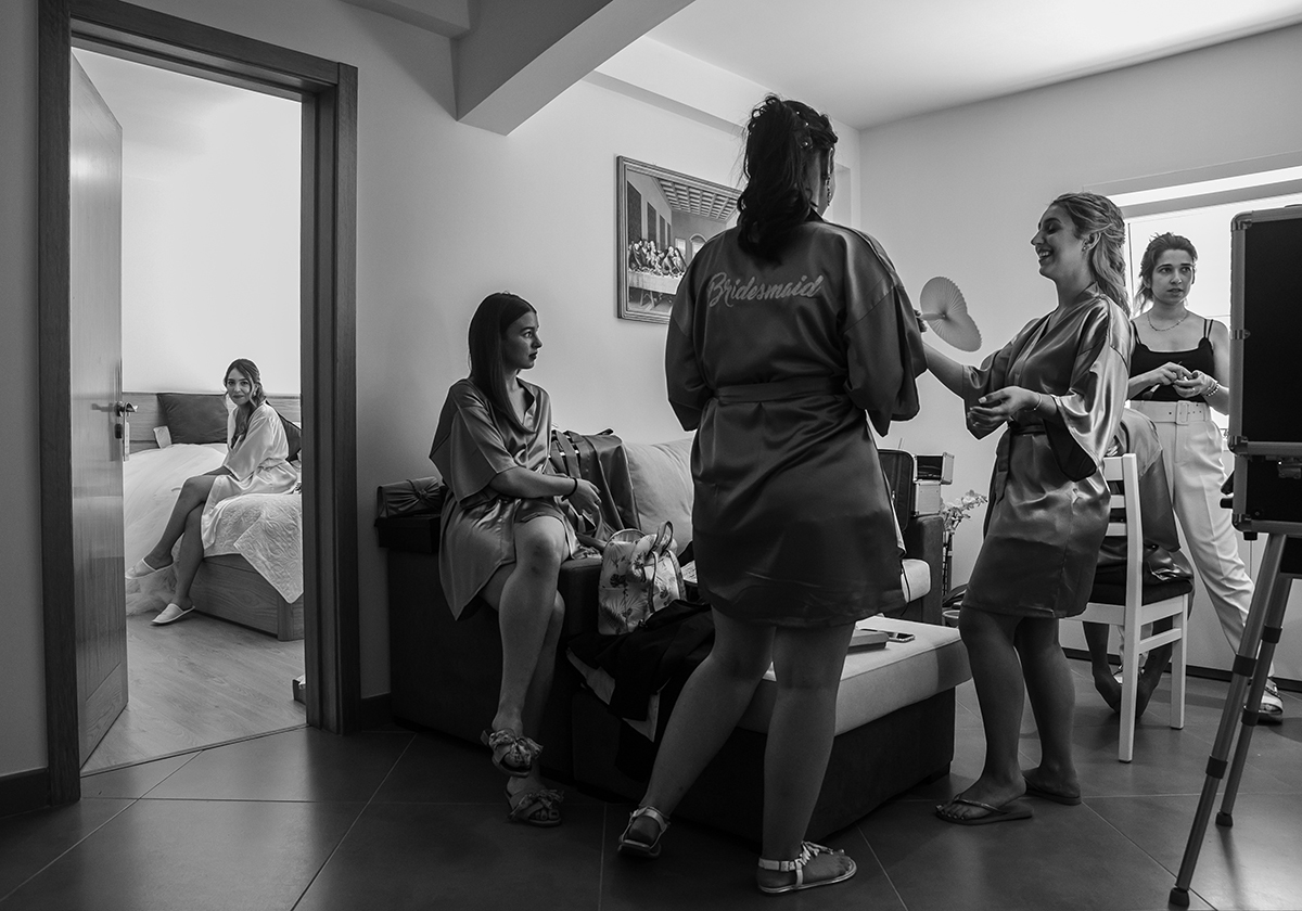 Um instante capturado entre risos e silêncios: de um lado, as madrinhas trocam palavras e gargalhadas, uma delas com um robe a dizer “Bridesmaid”; do outro, Patrícia, sentada na cama, olha para a câmara com serenidade e um sorriso cheio de verdade.