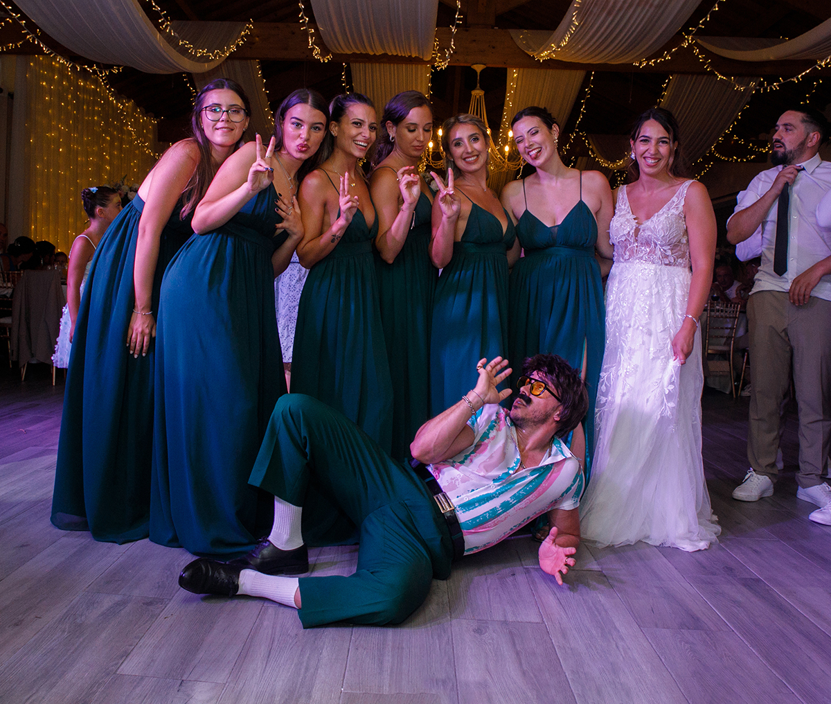 Foto de grupo com a noiva e as amigas, todas a sorrir para a câmara com dois dedos no ar. O Sr. Camaleão está deitado no chão, com uma mão pousada e a outra levantada, a dar ainda mais energia ao momento.