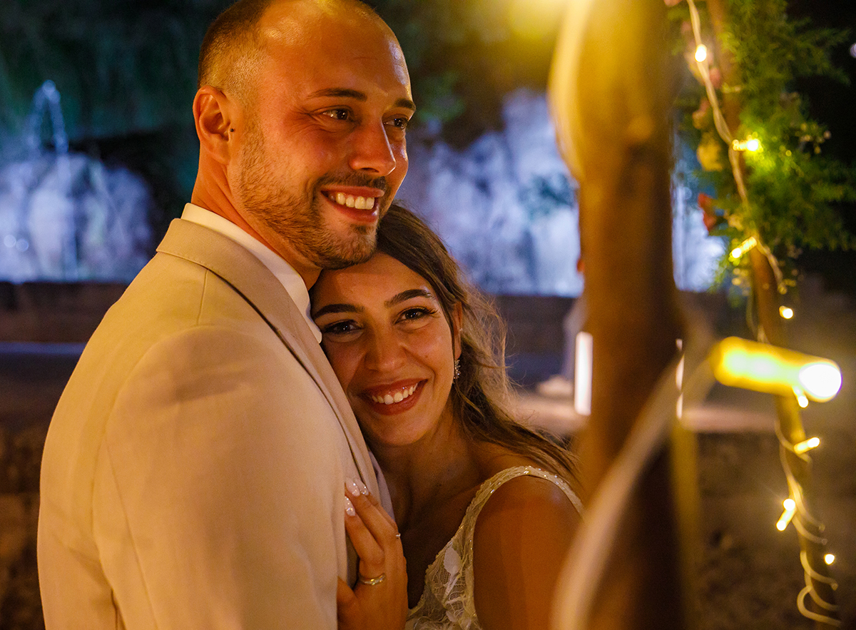 Um plano mais próximo dos noivos, do peito para cima. Ela encostada ao peito dele, com a mão sobre o coração dele. Ambos sorriem enquanto olham para o lado. À direita da imagem, vê-se um fio com luz que completa a composição com um detalhe delicado.