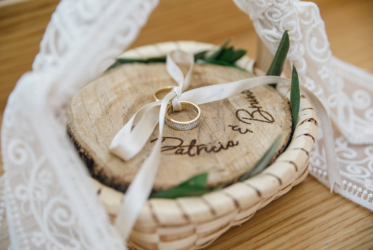 Num delicado plano de detalhe, um cesto adornado com carinho segura uma pequena placa em madeira com os nomes dos noivos gravados. Sobre ela, repousam duas alianças presas por lacinhos, promessas eternas embaladas com amor.