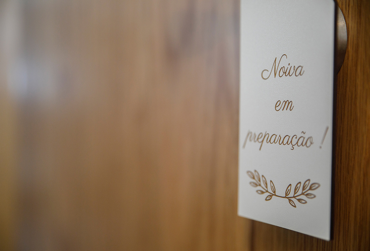 A porta de madeira, antiga e imponente, aparece desfocada ao fundo, mas o olhar é imediatamente atraído pela placa pendurada no puxador que anuncia “Noiva em Preparação!”. A madeira e o metal envelhecido parecem refletir a tradição.