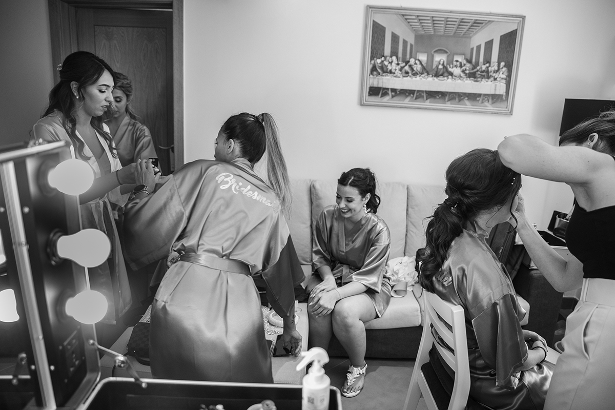 Em preto e branco, o quarto revela uma cena íntima e descontraída, onde a noiva e as suas amigas estão imersas na arte da maquilhagem, num momento de diversão despreocupada, quase como se o tempo tivesse parado ali, apenas para elas.