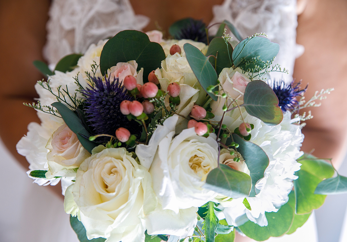 Flores brancas, roxas e rosa entrelaçam-se num bouquet que exala romantismo. Atrás dele, desfocada mas presente, Patrícia observa o início de um novo capítulo.