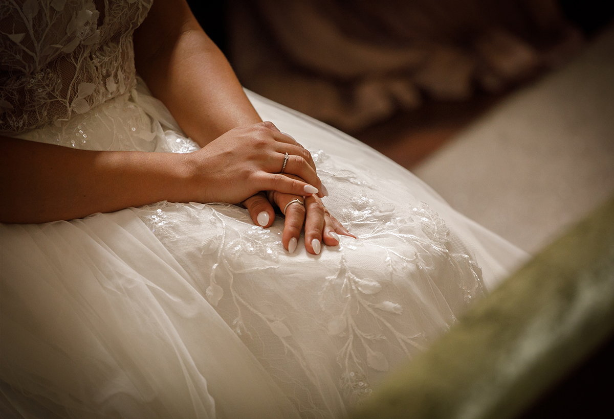As mãos da noiva repousam delicadamente sobre o seu vestido adornado. O plano com vinheta e detalhe celebra a beleza dos pequenos gestos.