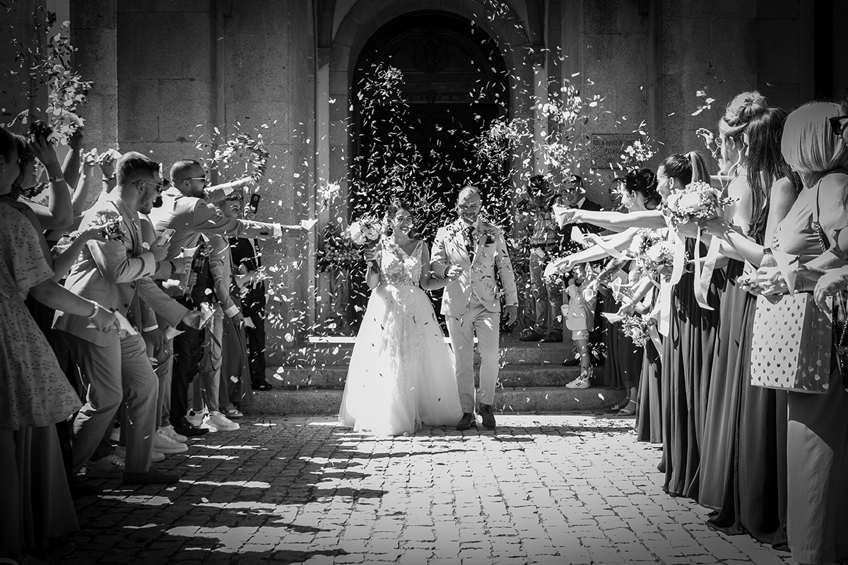 Em preto e branco, os noivos saem da igreja entre flores lançadas. Cabeças baixas, sorrisos tímidos, um momento de beleza natural e genuína.
