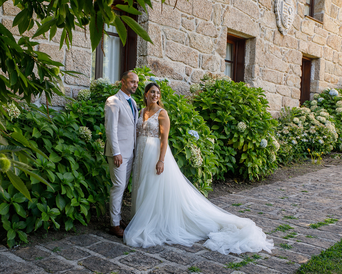 Num jardim em frente a uma casa antiga, os noivos caminham lado a lado. O vestido estende-se sobre as pedras como um rio de luz.