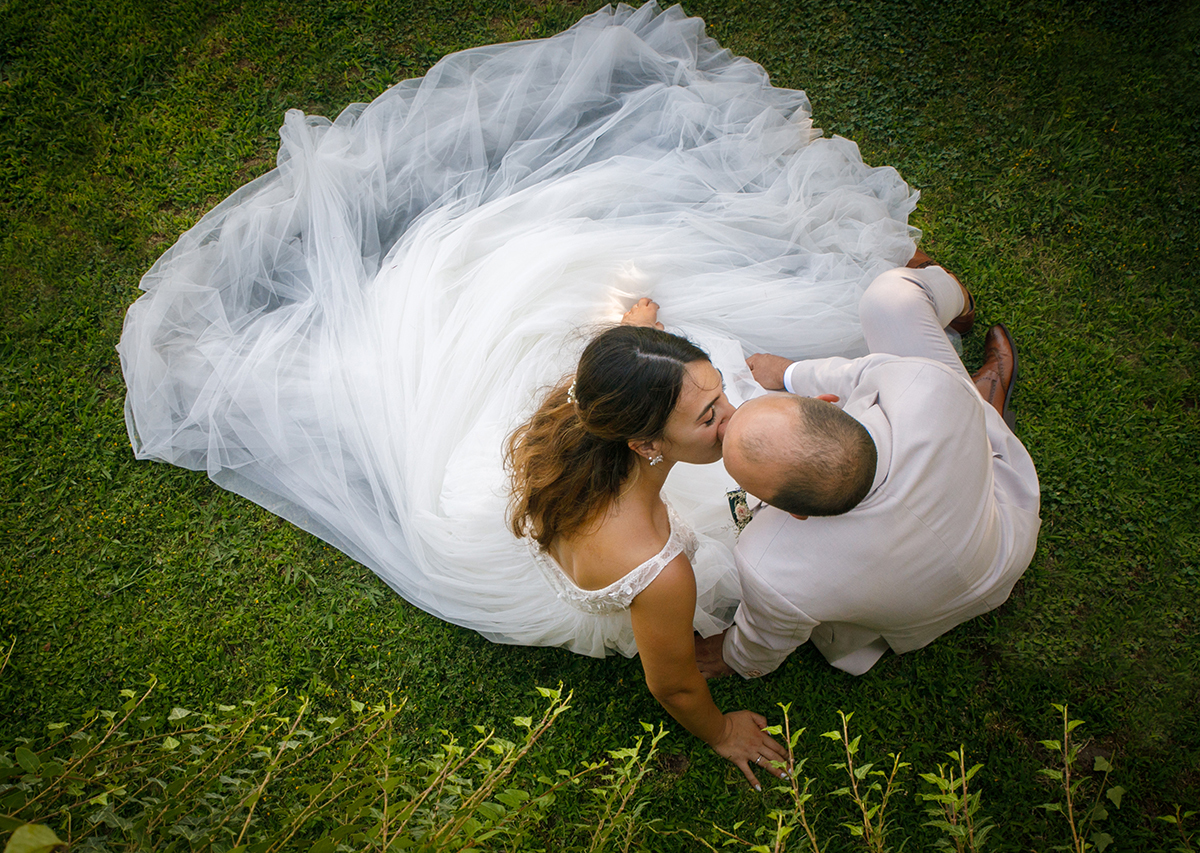 Sentados na relva, vistos de cima, os noivos beijam-se com a doçura de quem encontrou um lar no outro.