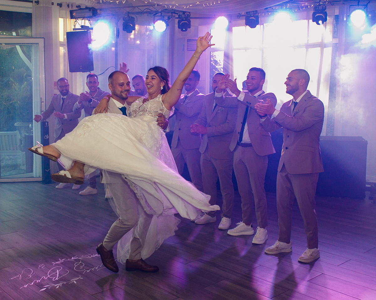 O noivo leva a noiva ao colo, as luzes da festa pintam o cenário, e os amigos aplaudem, um momento de pura alegria cinematográfica.