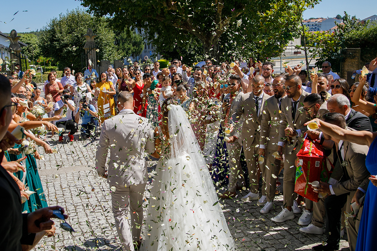 De costas, os noivos percorrem um caminho entre convidados que lançam folhas e flores ao ar. O sol brilha, como se abençoasse cada passo.