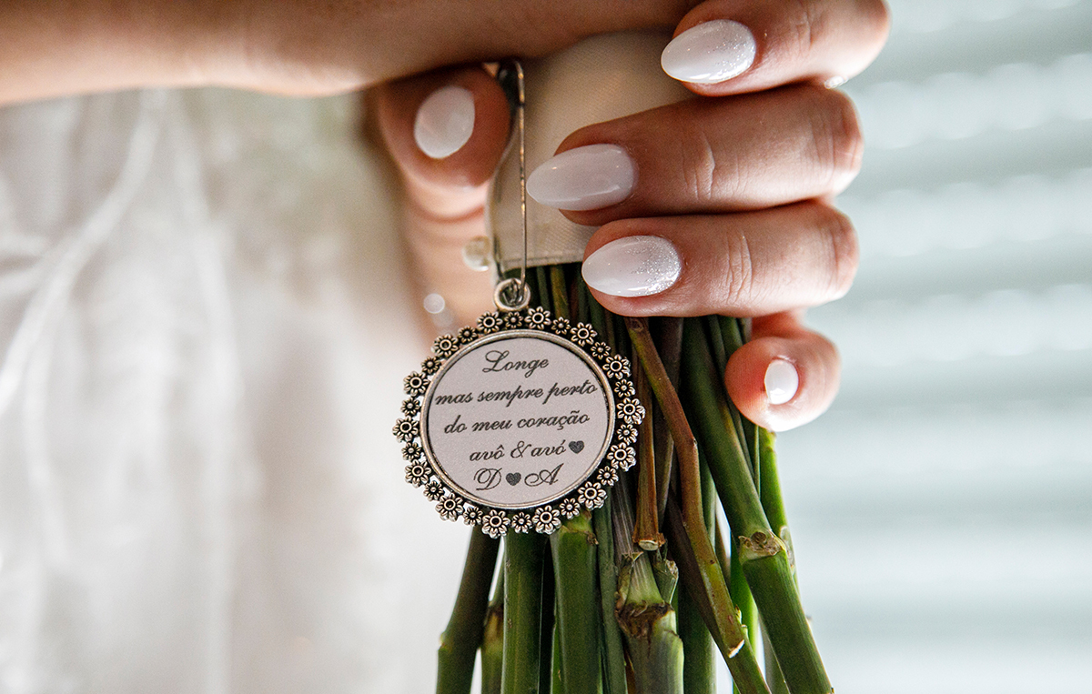 Um plano comovente mostra as mãos da noiva sobre o vestido. O bouquet descansa, e entre os pés flores, uma placa diz: 