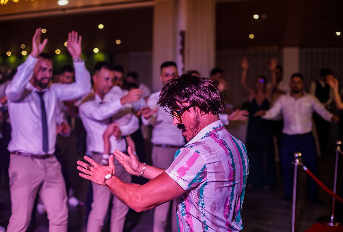 No centro da pista, o Sr. Camaleão dança com entusiasmo, enquanto os convidados, desfocados ao fundo, o seguem em ritmo e espírito.