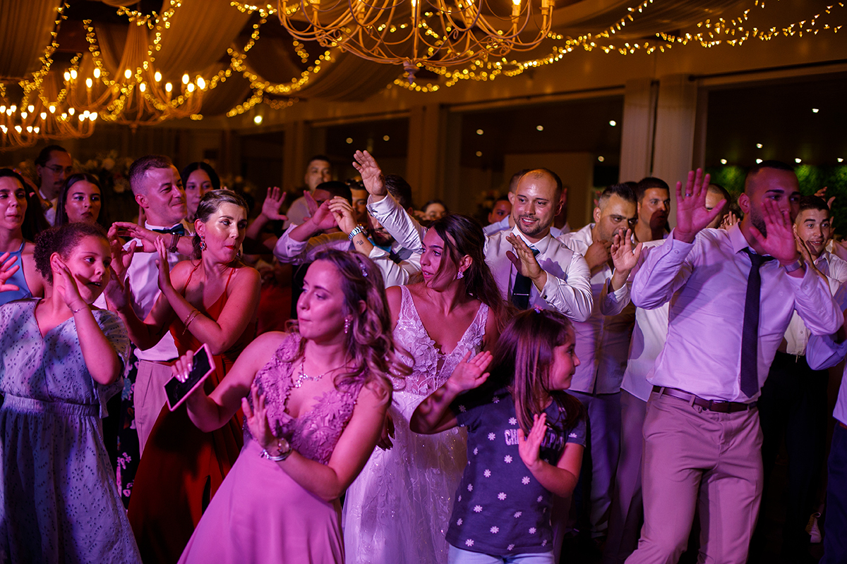 Os convidados aparecem cobertos por uma luz rosa, a dançar com as mãos viradas para cima e para o lado, num momento cheio de cor e movimento.