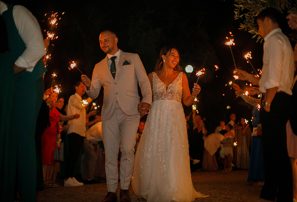 Nesta imagem, os noivos estão no centro num ângulo ligeiramente contrapicado. Ambos com sparkles nas mãos, sorriem para os convidados enquanto a luz das faíscas ilumina a cena.