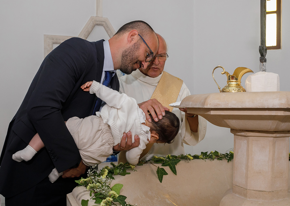 O padrinho inclina Francisco sobre a Pia Batismal enquanto o padre derrama a água benta na sua cabeça. As flores delicadas sobre a pedra completam o cenário sagrado e puro.