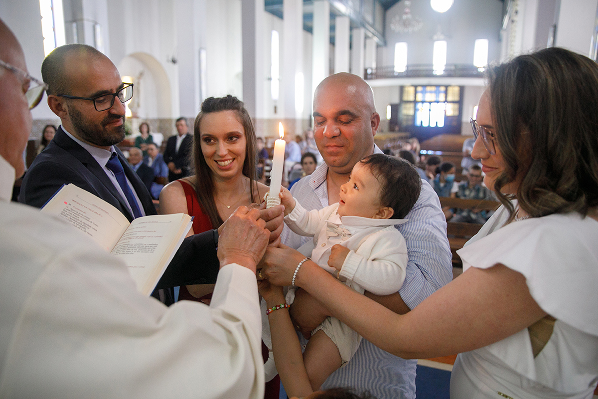 Reunidos na igreja, o padre lê em voz alta enquanto os pais e padrinhos seguram velas. O pai, com Francisco ao colo, partilha esse instante de fé e união em família.