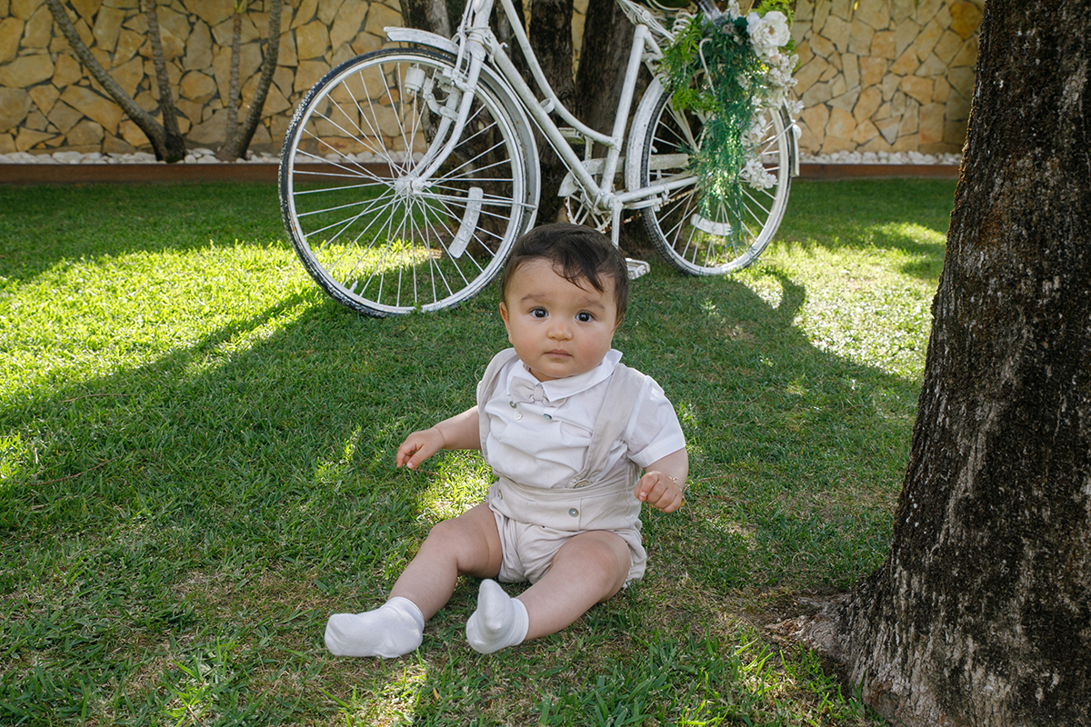 Junto ao tronco de uma árvore, Francisco ergue as mãos e olha para a câmara com seriedade. Ao fundo, a bicicleta branca decorada com flores acrescenta um toque de sonho ao retrato.