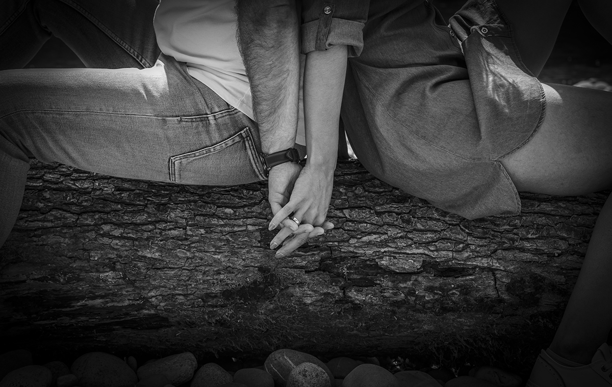 As mãos entrelaçadas de Marta e Mathieu ganham destaque. A textura da madeira do tronco onde se sentam enriquece o plano, que foca os gestos simples e íntimos do casal. Sessao Solteiros na Suiça