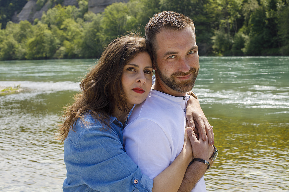 Marta está a abraçar Mathieu por trás, pousando delicadamente as mãos sobre o peito dele, formando um coração. Mathieu, com um sorriso suave, coloca as mãos por cima das dela. Marta mantém uma expressão serena, mas transmite felicidade. 