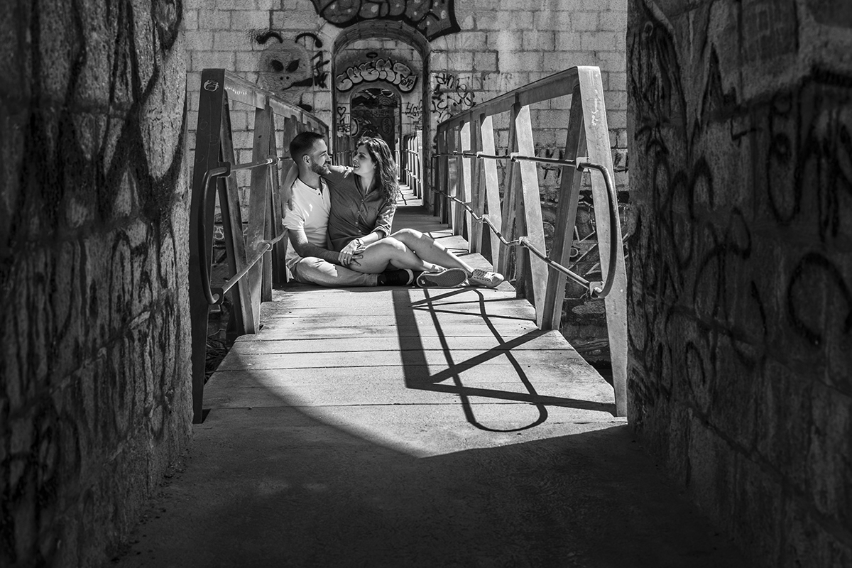 Os dois estão sentados no meio de uma ponte, abraçados. Olham um para o outro com ternura. A estrutura antiga da ponte e o cenário à volta acrescentam um toque romântico e nostálgico ao momento.