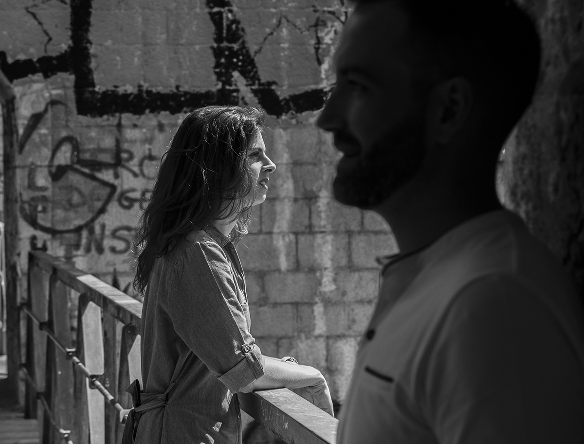 Numa aproximação mais íntima, Mathieu encosta-se a uma parede com expressão serena. Marta, ao lado, apoia-se no varão da escada, olhando para o horizonte com um leve sorriso. Um momento de introspeção partilhado.