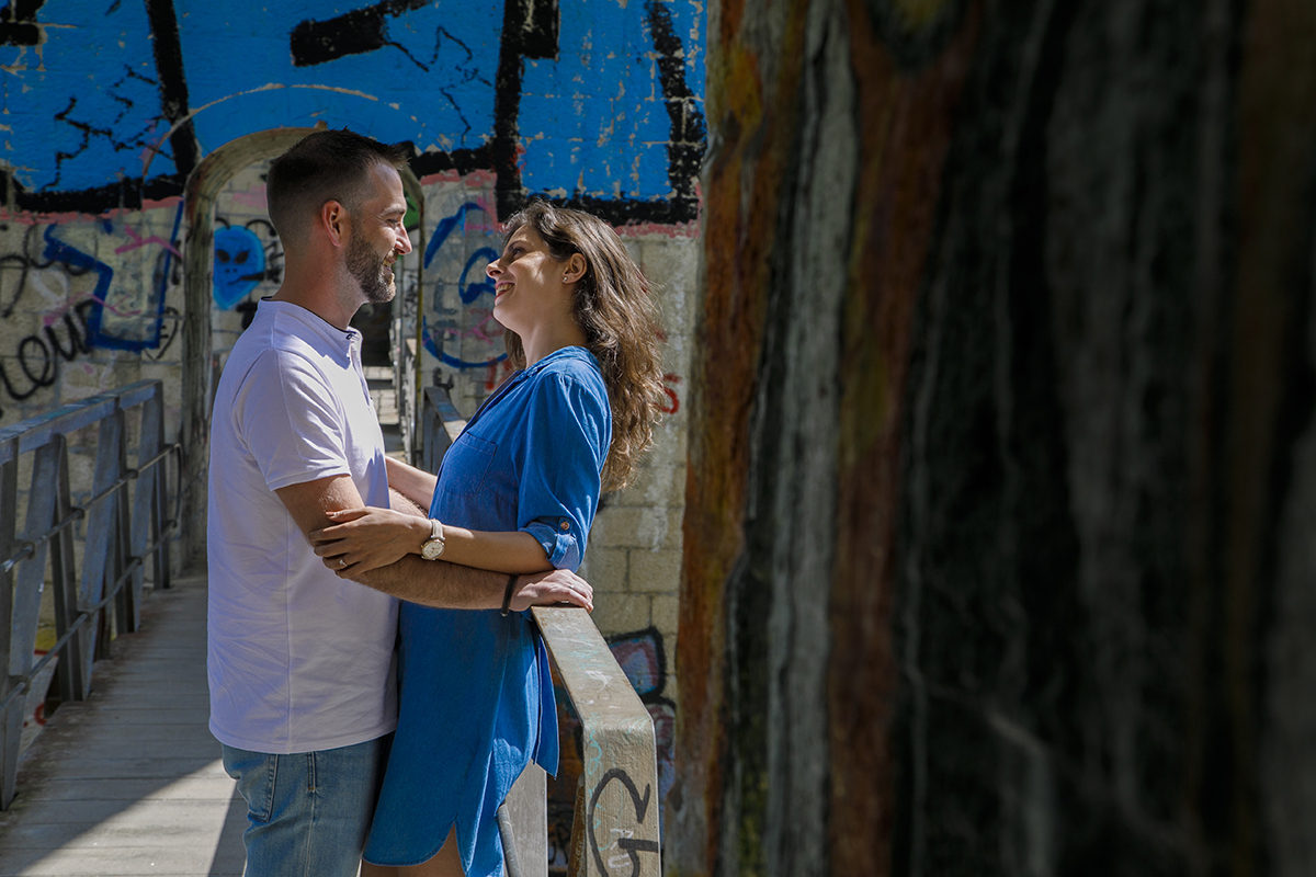 Marta está encostada à grade da ponte, Mathieu à sua frente com as mãos no corrimão. Os dois trocam olhares doces. Ao fundo, um grafitti azul na parede combina com o vestido de Marta, criando harmonia na composição visual.