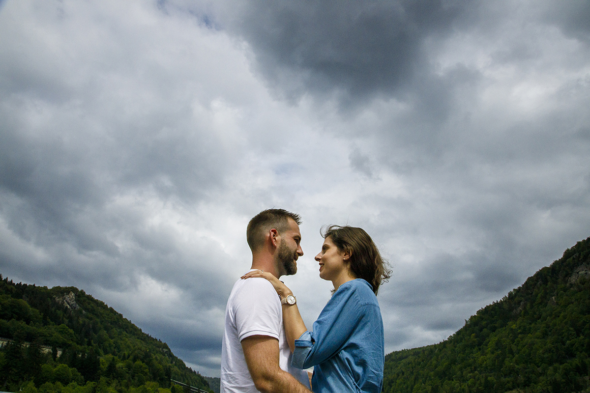 Num ângulo ligeiramente contrapicado, vemos o casal da cintura para cima. Marta apoia a mão no ombro de Mathieu, e ambos trocam olhares profundos e pensativos. Ao fundo, montanhas verdes e um céu dramático criam um ambiente inspirador.