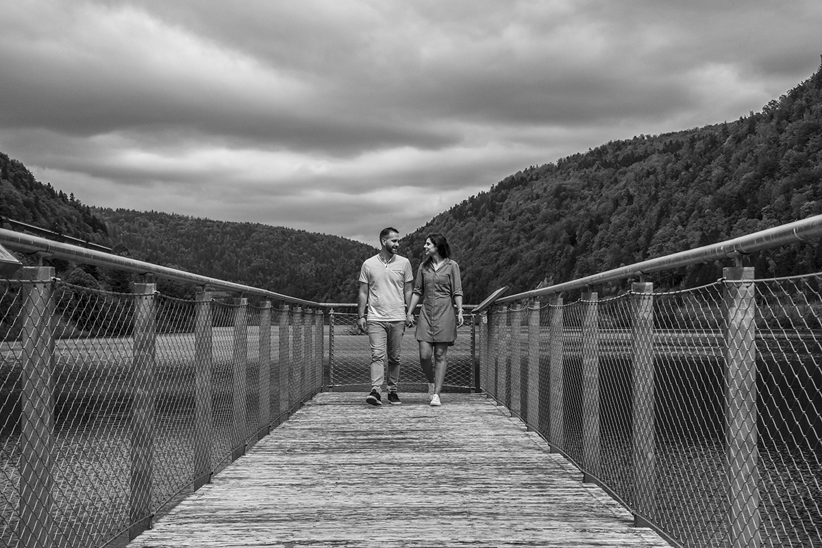 A preto e branco, os dois caminham de mãos dadas sobre uma ponte entre montanhas. Sorriem um para o outro, envoltos pela paisagem imponente e o amor que partilham.