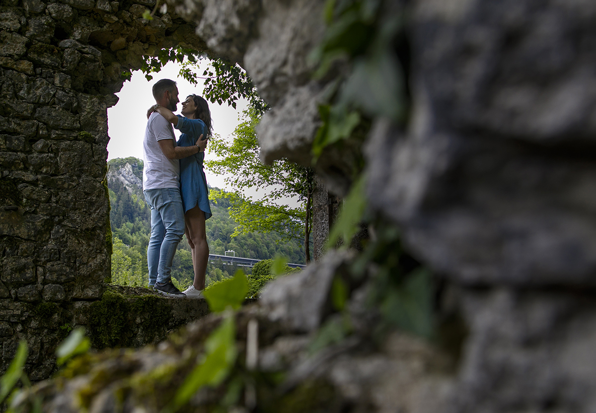 Sentados no parapeito da janela, abraçam-se e trocam um olhar apaixonado. No lado direito da imagem, uma parede com trepadeiras desfocada adiciona textura e interesse à composição.