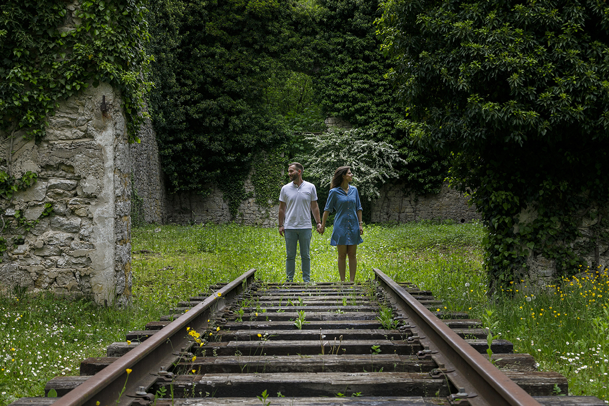 Num cenário rústico com linhas de comboio cobertas de verde, o casal está no fim da linha, de mãos dadas. Cada um olha para um lado diferente, num momento de reflexão envolto em natureza.