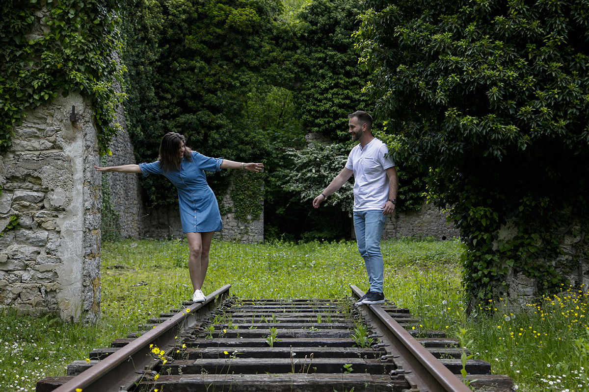 Ambos caminham sobre os trilhos do comboio. Marta tenta equilibrar-se na viga de ferro com os braços abertos, e Mathieu observa-a atento, com a mão estendida, pronto para a amparar se necessário.