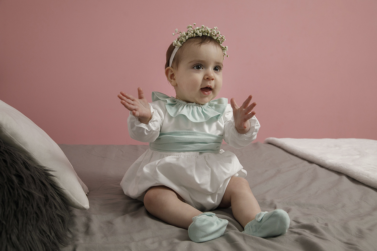 Ainda sobre a cama, Valentina bate palmas e olha de lado. A sua expressão séria e feliz revela a mistura de descoberta e encanto da infância.