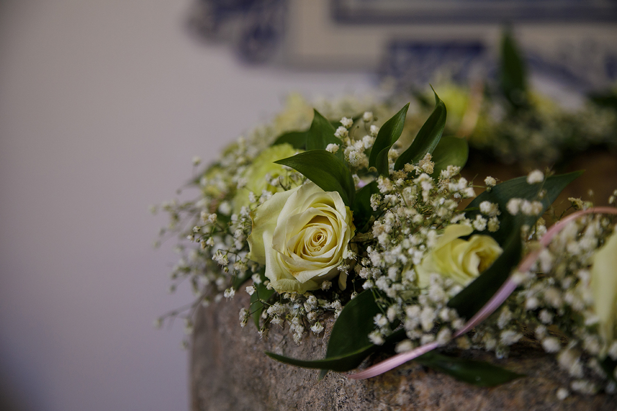 Num detalhe encantador, a Pia Batismal de pedra é adornada com verdes, pequenas flores brancas e amarelas, e uma fita rosa. Um altar natural para um momento sagrado.