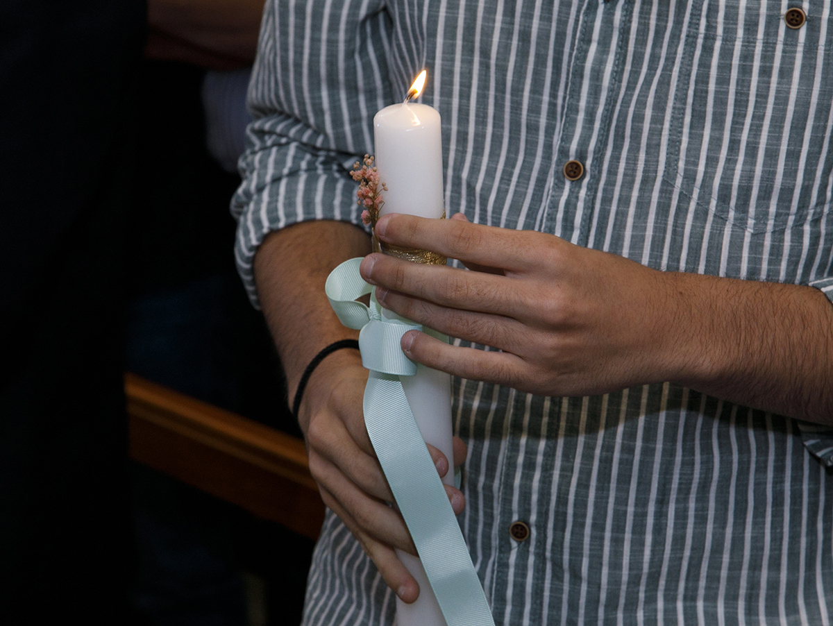 O padrinho segura delicadamente a vela do batismo. O laço verde combina com a sua camisa. Um detalhe simbólico que une o gesto à emoção.