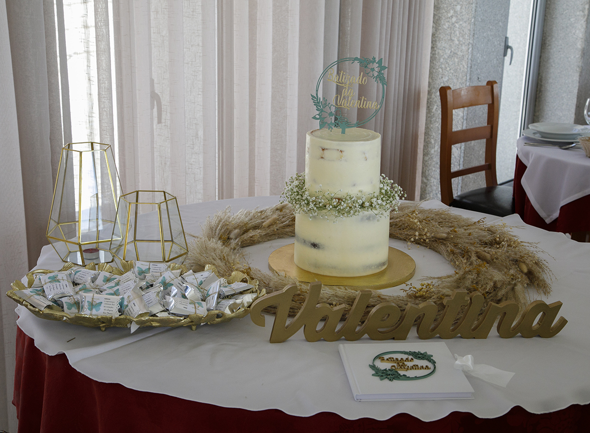 A mesa do bolo é um altar de doces e significados. O bolo branco de dois andares traz flores entre os níveis e um topo a dizer Batizado da Valentina. À volta, trigo, uma placa com o nome e um cenário de doçura.