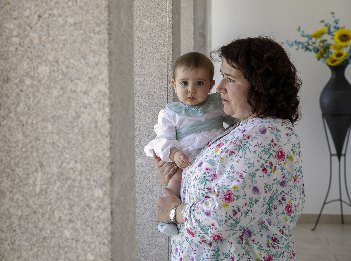 Entre pilares e janelas, Valentina está ao colo da mãe, que olha para a janela. Ela, por sua vez, encara a câmara com olhos atentos. Atrás, um vaso de girassóis emoldura a cena com luz.
