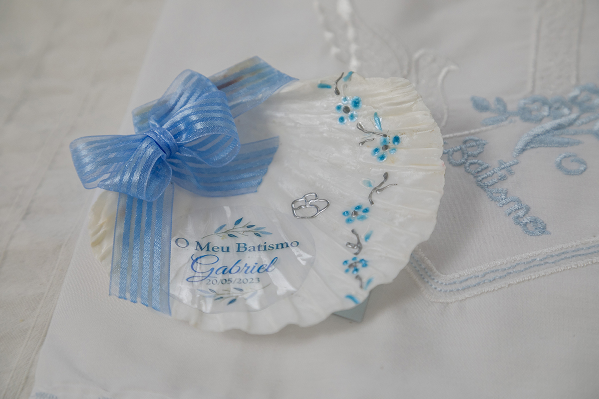 Num plano de detalhe repousa a concha branca, adornada com flores e espigas douradas, onde se lê, em letras delicadas, “O Meu Batizado Gabriel”. Um laço azul pousa no canto. Por baixo, o pano branco com bordado simples diz “Batismo