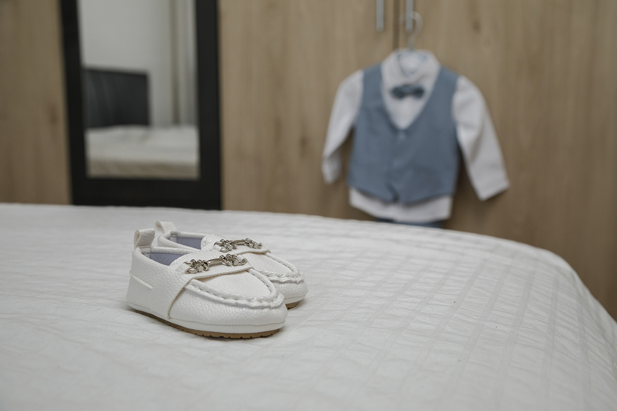 Sobre a colcha branca imaculada, repousam pequenos sapatinhos brancos, como se aguardassem o início de uma nova caminhada. Ao fundo, um roupeiro de madeira com espelho reflete o ambiente, enquanto o fato branco e azul do Gabriel está pendurado no roupeiro