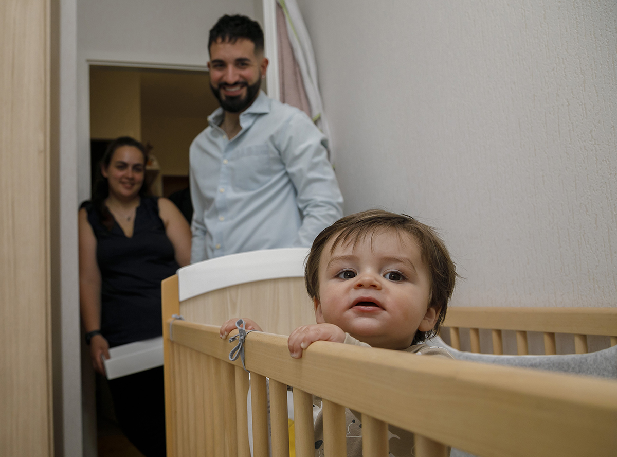 Junto à porta do quarto, a mãe sorri com uma caixa branca nos braços. O pai, mais à frente, olha para Gabriel com admiração. O pequeno encosta-se à sua caminha, com as mãozinhas no corrimão e o olhar doce ligeiramente desviado.