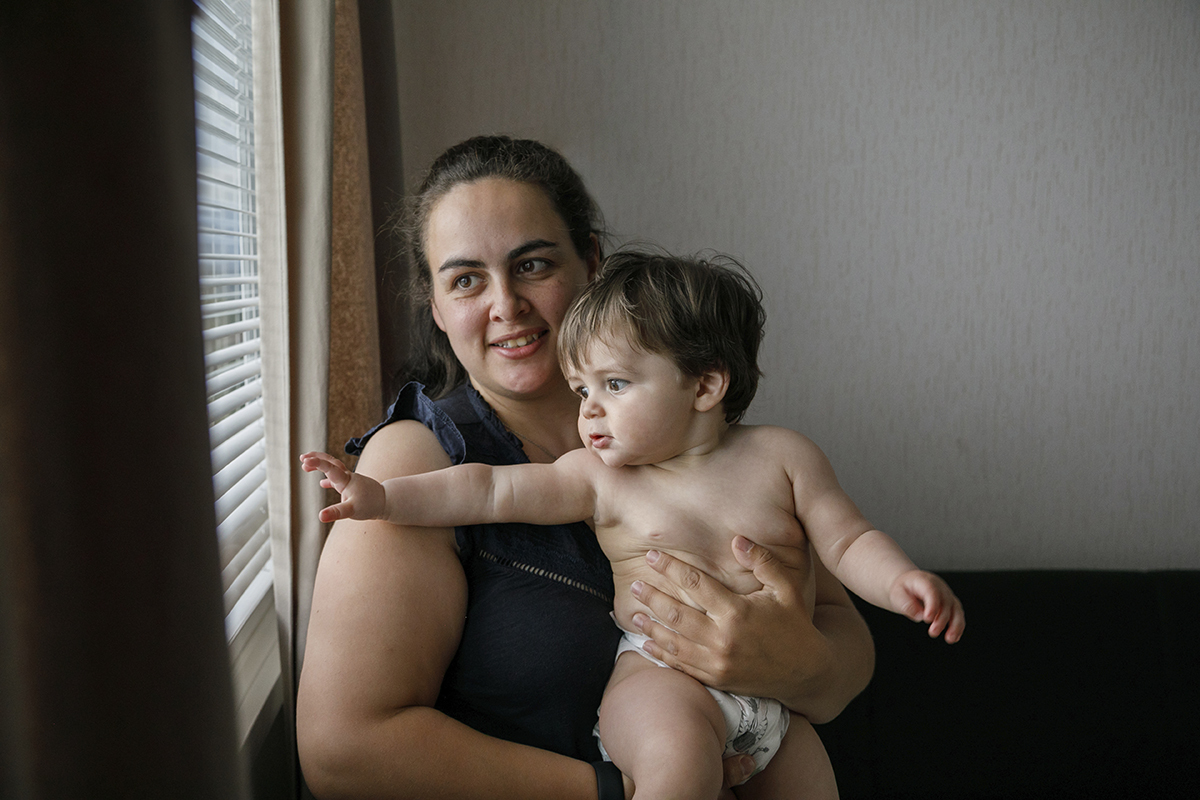 Junto à janela, o colo da mãe torna-se refúgio. A luz banha os dois, e o pequeno Gabriel encosta a mão no vidro, contemplando o mundo. Ambos sorriem para a claridade, numa cumplicidade suave entre mãe e filho.