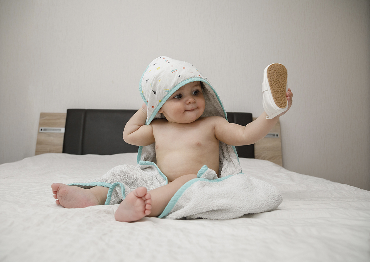 Sentado sobre a cama, com a manta branca a envolvê-lo da cabeça aos pés, Gabriel segura o sapatinho com uma mão e com a outra a ponta da manta que lhe cobre a cabeça. A cabeceira escura contrasta com a leveza do instante.