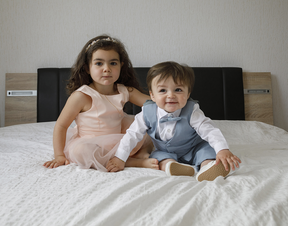 Na cama, lado a lado, os irmãos posam para a câmara. Ela, de vestido rosa, pousa a mão sobre a colcha, os pés encostados às pernas do irmão. Ele segura o sapatinho com uma mão e o vestido da irmã com a outra. Sérios, mas com alegria contida no olhar.