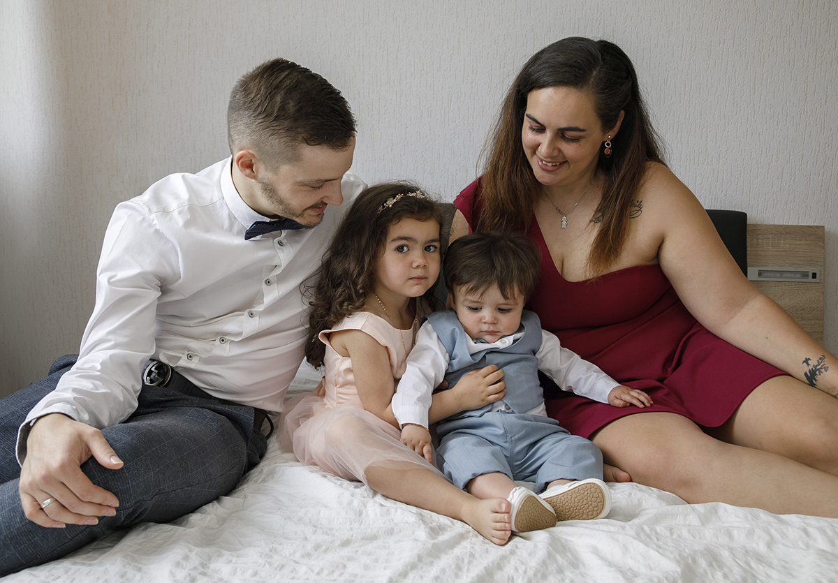 Agora mais próximos, os pais inclinam-se e sorriem para os filhos. A irmã olha diretamente para a lente, séria, enquanto Gabriel baixa os olhos com calma. Uma imagem de ternura e proteção.