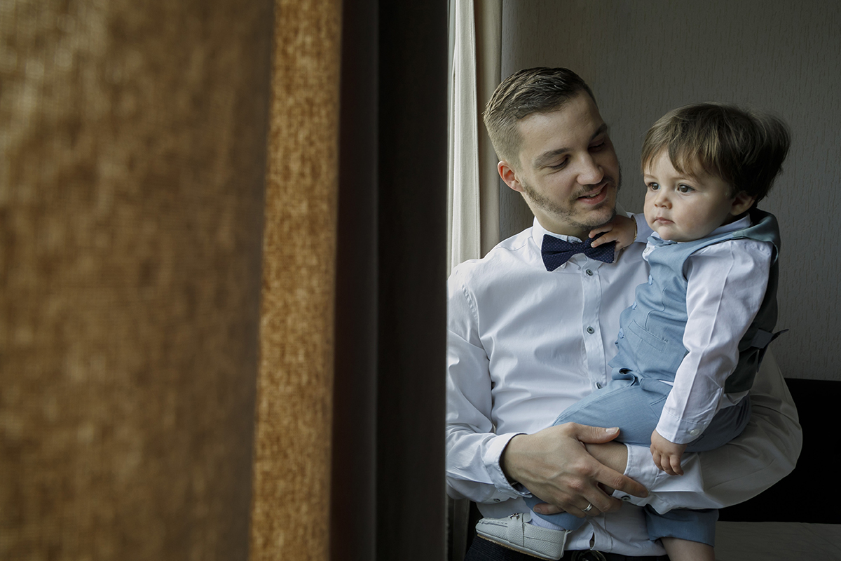 Encostado à janela, o pai segura Gabriel nos braços. O menino observa o exterior com olhar sereno enquanto o pai o contempla com carinho, um momento de silêncio entre gerações.